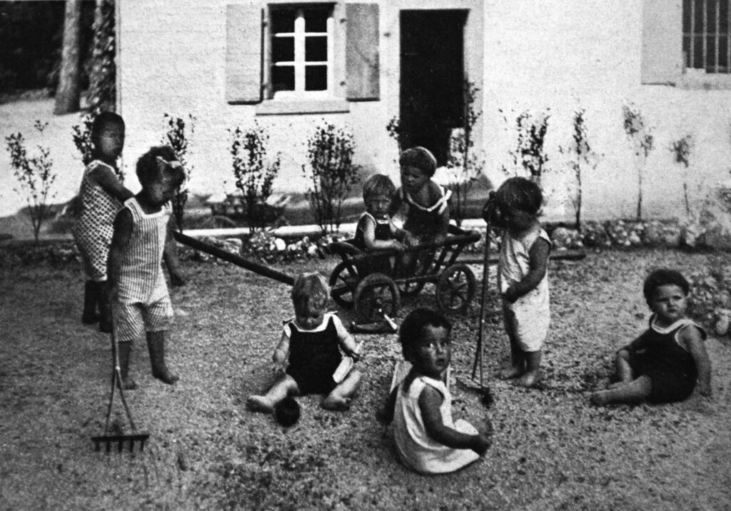 Kleinkinder spielen mit Gartenwerkzeug auf dem Boden vor dem Haus.
